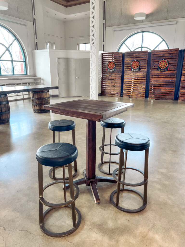 Wooden cocktail table with metal barstools in front of dartboard backdrops and whiskey barrel tables inside a modern industrial venue by In The Event.