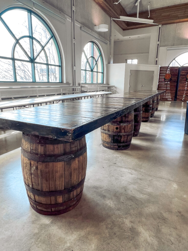 Long rustic event table supported by whiskey barrels beneath arched windows in an industrial venue designed by In The Event.