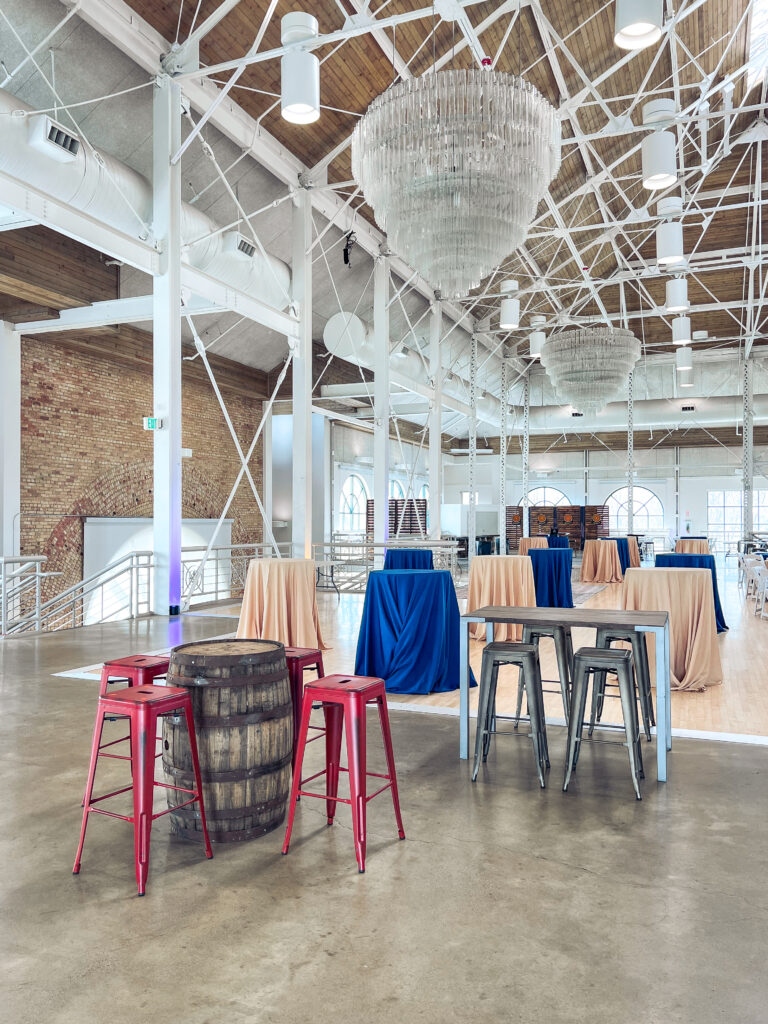 Spacious event venue featuring chandeliers, blue and tan cocktail tables, metal barstools, whiskey barrel tables, and leather lounge seating styled by In The Event.