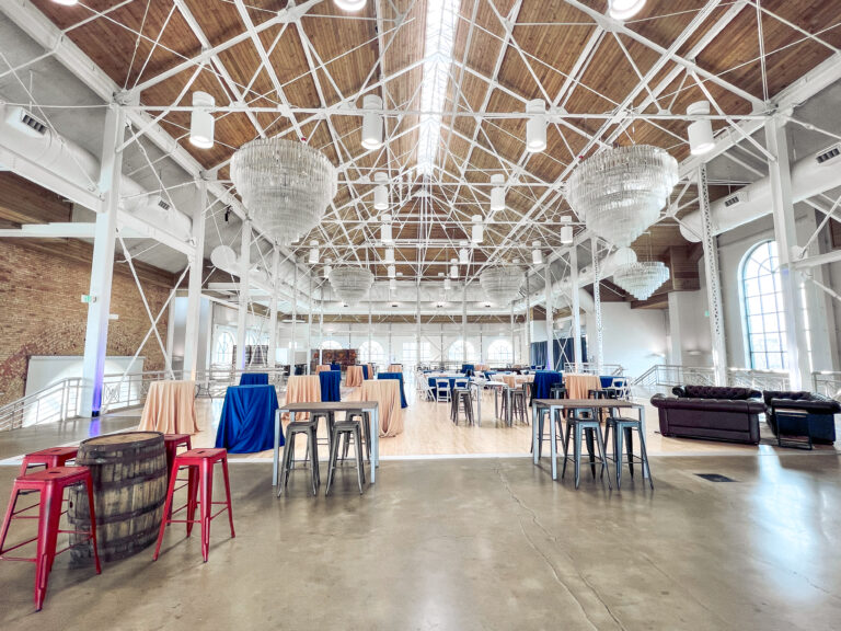 Spacious event venue featuring chandeliers, blue and tan cocktail tables, metal barstools, whiskey barrel tables, and leather lounge seating styled by In The Event.