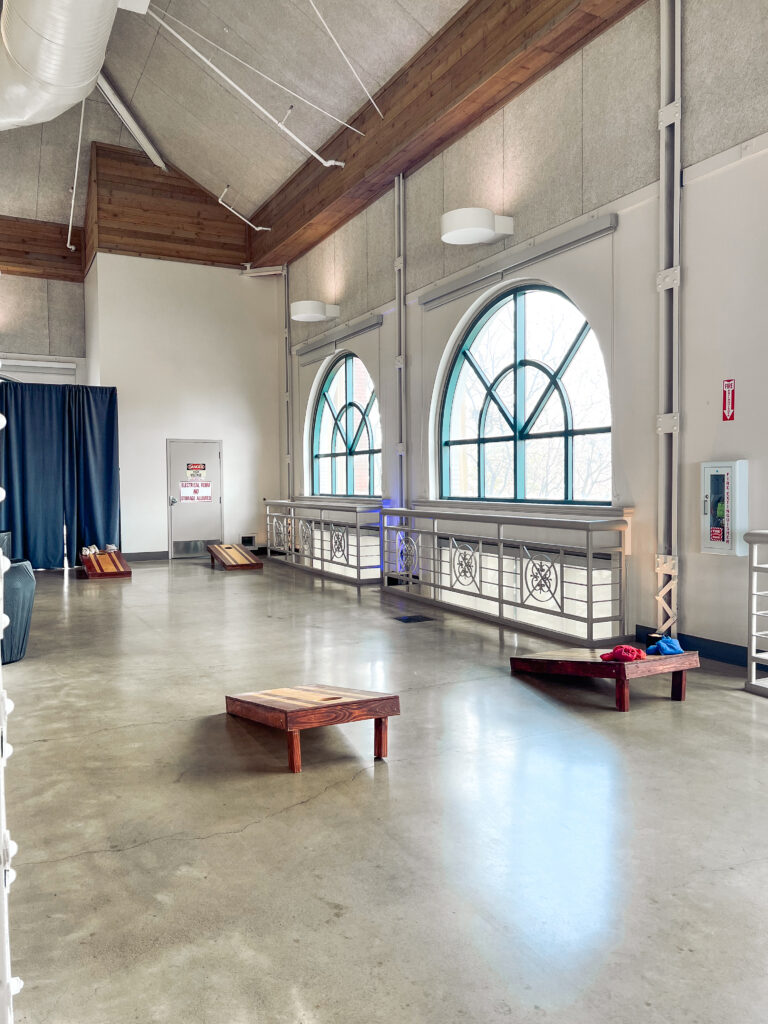 Bright event space with arched windows and polished floors featuring multiple wooden cornhole boards and colorful bean bags by In The Event.