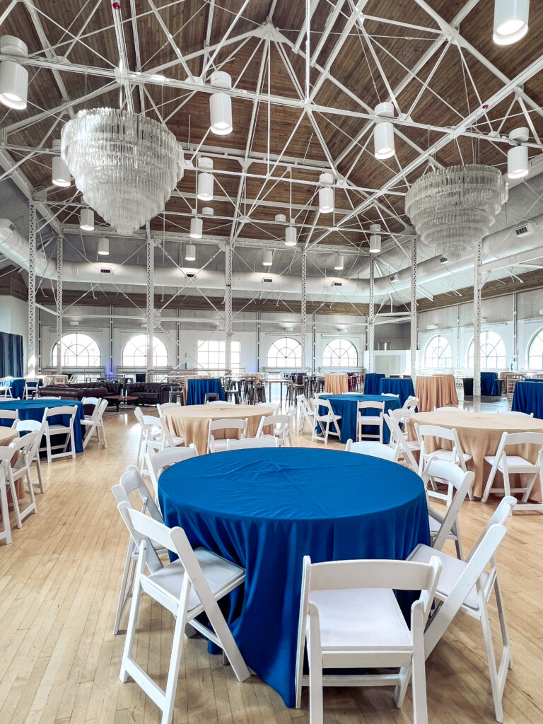 Elegant event venue with round tables covered in blue and tan linens, white chairs, and crystal chandeliers styled by In The Event.
