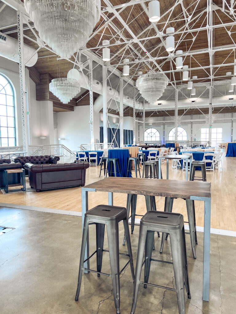 Open event venue with crystal chandeliers, blue and tan linens, and industrial-style metal bar tables and stools arranged by In The Event.