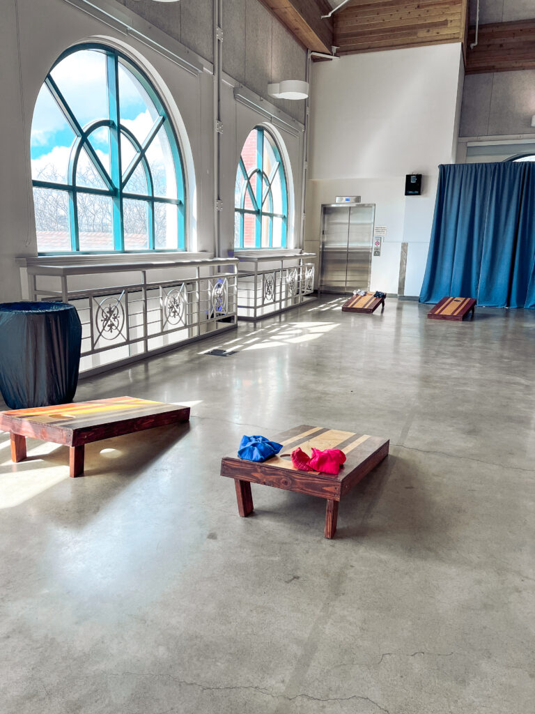 Custom wooden cornhole boards with red and blue bean bags arranged inside a bright event venue with arched windows and polished concrete floors.