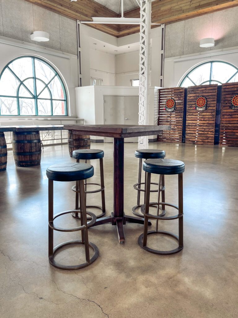 Rustic cocktail table with round metal barstools set in a bright venue featuring wood panel dartboard walls and barrel decor by In The Event.