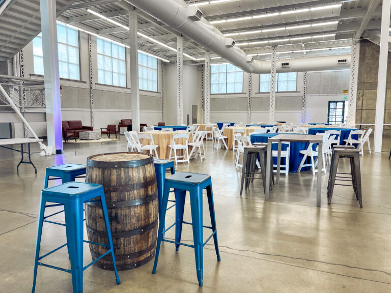 Spacious corporate event setup with cocktail tables, blue and tan linens, metal barstools, and whiskey barrel accents arranged by In The Event.