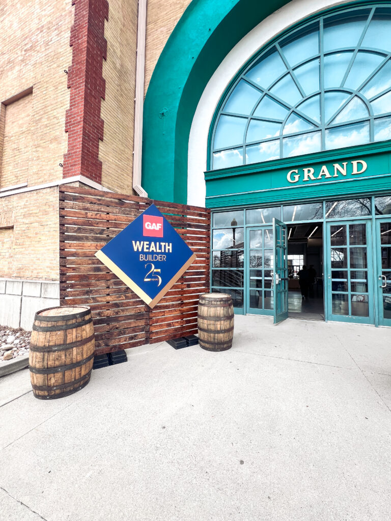 Rustic wood panel event backdrop with GAF Wealth Builder 25th Anniversary signage and whiskey barrel decor outside The Grand venue entrance.