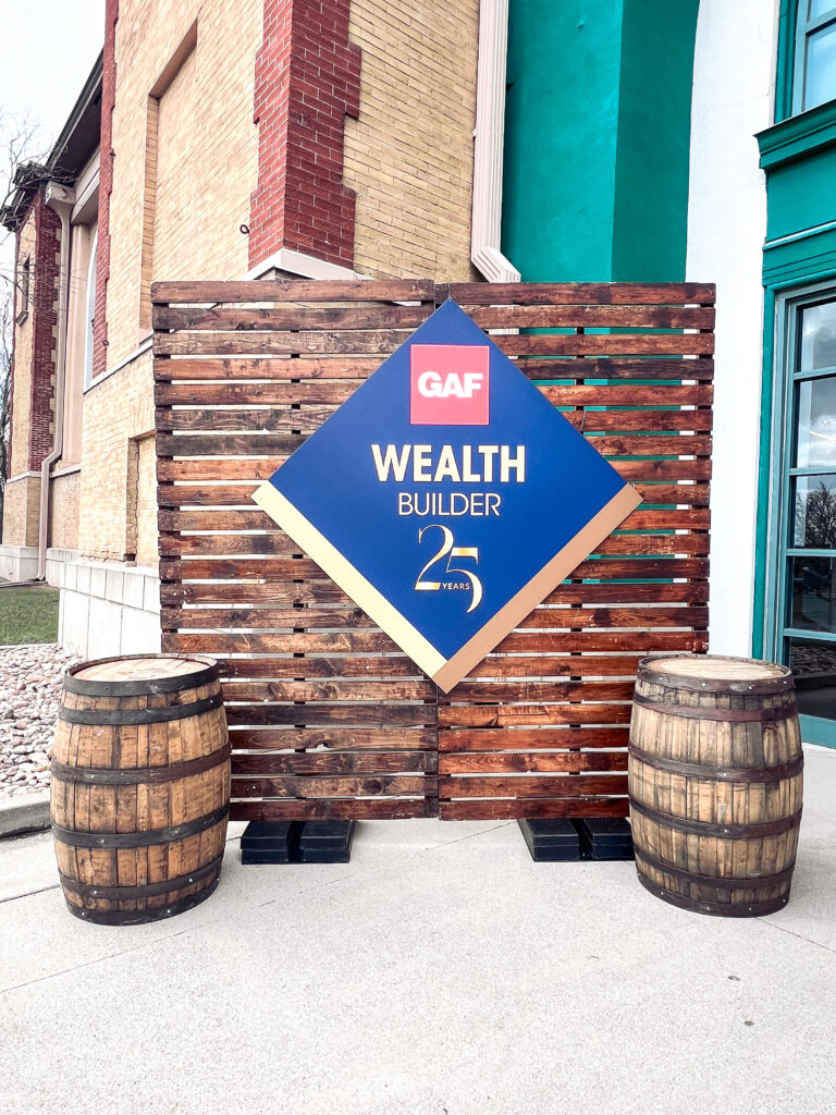 Outdoor event setup featuring a wooden backdrop with a large GAF Wealth Builder 25 Years sign flanked by two rustic whiskey barrels.