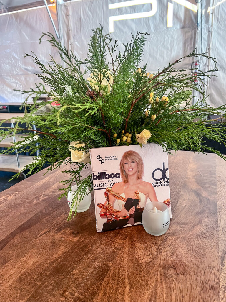 Creative floral arrangement in a vase wrapped with a Billboard Music Awards magazine cover, featuring greenery, white roses, and berry accents on a wooden table.