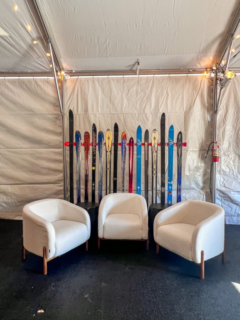 Three modern white lounge chairs arranged in front of a creative backdrop made from vintage skis inside a tented event space with string lighting.