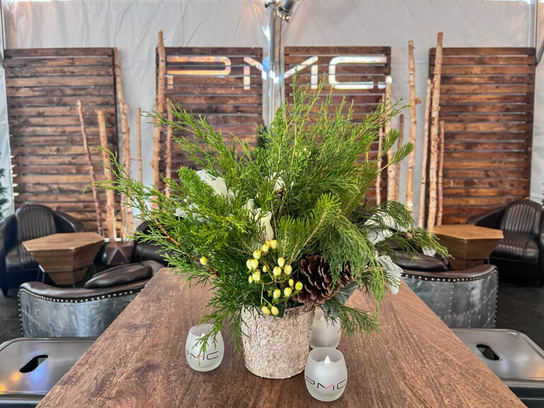 Close-up of a rustic floral arrangement featuring pinecones, white roses, evergreen branches, and yellow berries in a birch vase on a wooden table.