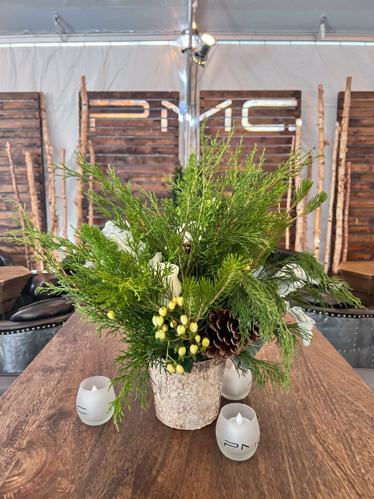 Close-up of a rustic floral arrangement featuring pinecones, white roses, evergreen branches, and yellow berries in a birch vase on a wooden table.