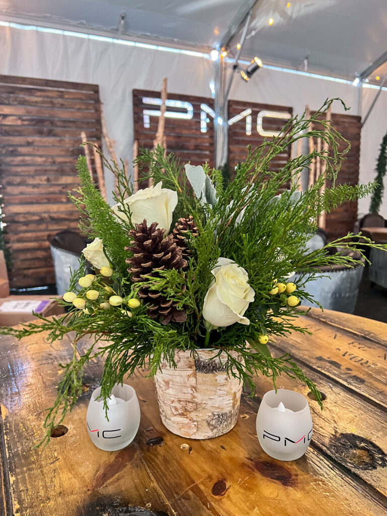 Close-up of a rustic floral arrangement featuring pinecones, white roses, evergreen branches, and yellow berries in a birch vase on a wooden table.