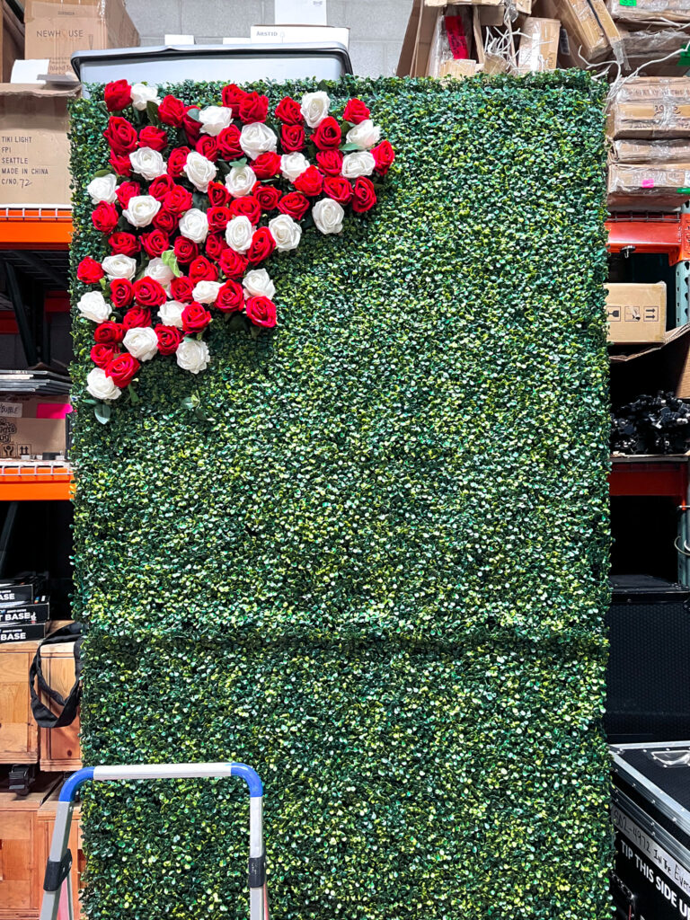 Custom greenery wall event backdrop featuring artificial boxwood hedge with red and white rose arrangement forming University of Utah U logo for graduation ceremony