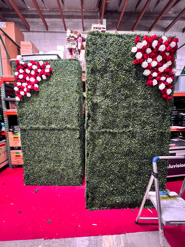 Modular event backdrop panels featuring artificial boxwood hedge walls with red and white rose arrangements forming University of Utah U logos for graduation ceremony photo wall