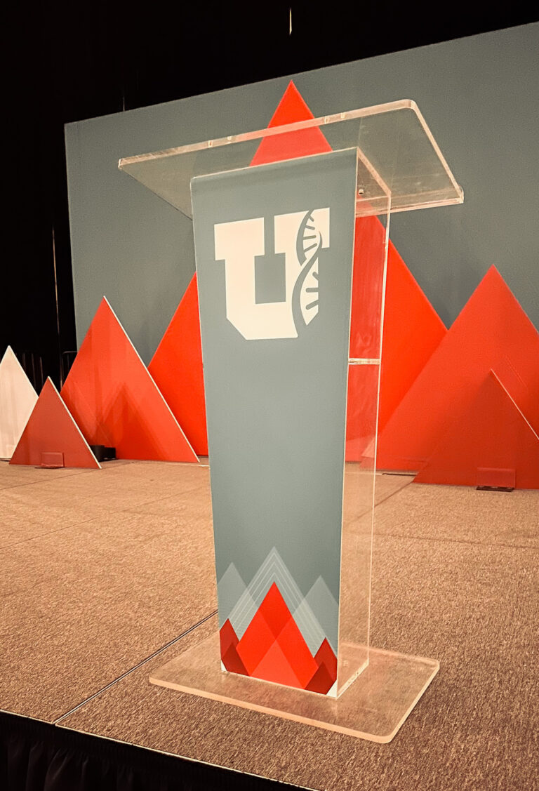 Clear acrylic podium with custom branding featuring a logo on the front, set on a stage rentals area with orange and white geometric event props in the background.