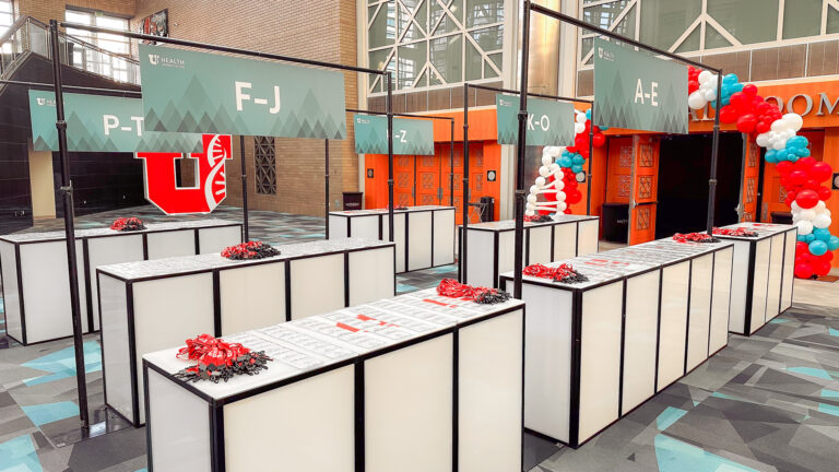 Long rows of white light-up LED furniture rentals registration desks with overhead lettered custom event signage, red lanyards, and branded backdrop elements for trade show logistics.