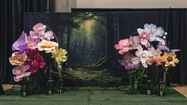 Decor backdrop featuring oversized paper flowers in pink, purple, and yellow surrounding a forest scene with lanterns and moss accents.