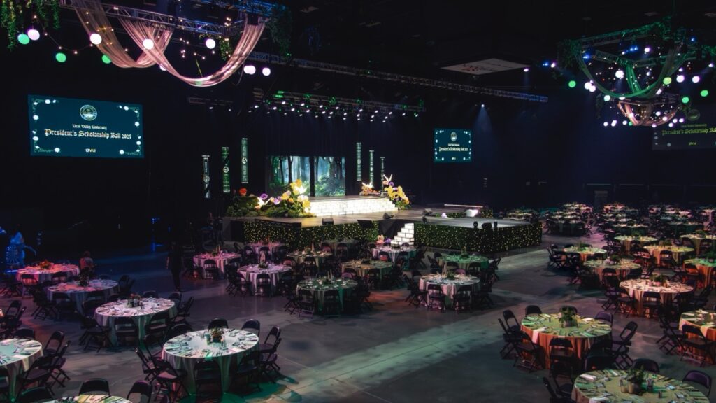 Wide view of the UVU President&rsquo;s Scholarship Ball 2025 featuring an illuminated stage, hanging greenery, and round guest tables arranged for the gala dinner.
