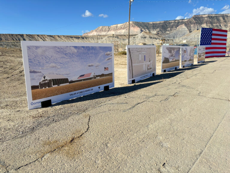 Architectural renderings of the Valar Atomics Ward 250 facility displayed outdoors along a paved area, with an American flag and mountain backdrop.