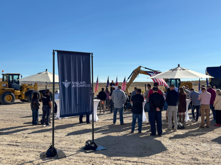 Guests attend the Valar Atomics groundbreaking ceremony outdoors with branded signage, American flags, and construction equipment in the background.