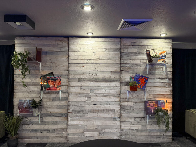 Custom event backdrop featuring whitewashed reclaimed wood panels with floating acrylic shelves displaying vintage magazines and greenery for rustic themed event decor