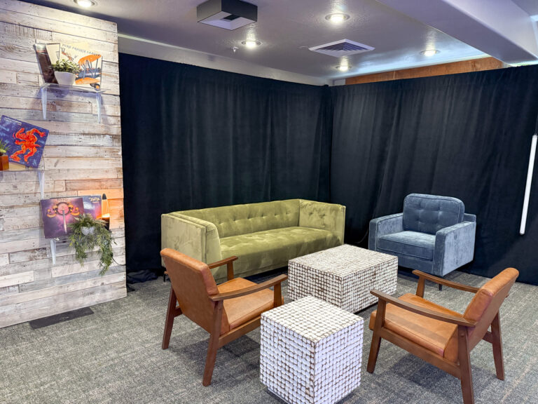 Event lounge furniture rentals featuring green velvet sofa, blue velvet chair, cognac leather armchairs, and mirrored cube ottomans with black pipe and drape backdrop and rustic wood wall