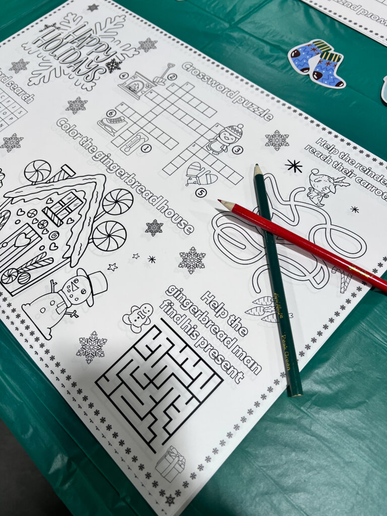 A festive craft table featuring a custom activity sheet with "Happy Holidays" puzzles, mazes, and coloring sections, used during the UA Local 140 event at the MAEC.