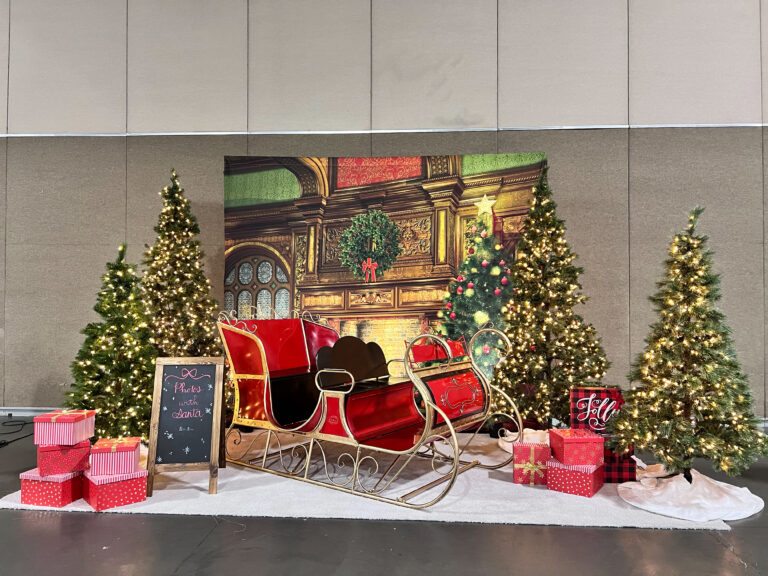 A festive photo op set featuring a classic red and gold Santa sleigh surrounded by wrapped gifts, four prelit pine trees, and a "Photos with Santa" chalk board sign positioned in front of a cozy holiday fireplace backdrop.