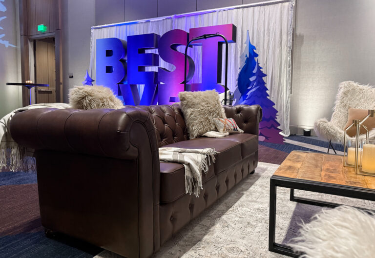 Brown leather Chesterfield sofa with faux fur and fringe throw pillows in front of a large illuminated "BEST" dimensional letter backdrop with blue and purple event lighting, winter tree cutouts, white pipe and drape, a faux fur accent chair, rustic wood coffee table with gold candle lanterns, and a gooseneck microphone stand at the Best Ever Conference at Hyatt Regency.