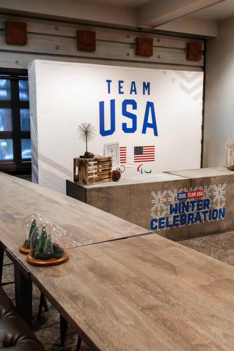 Team USA 2026 Winter Celebration event setup featuring a custom branded backdrop, rustic wood reception desk with pine cone decor, mini glass dome centerpieces, and American flags on display.