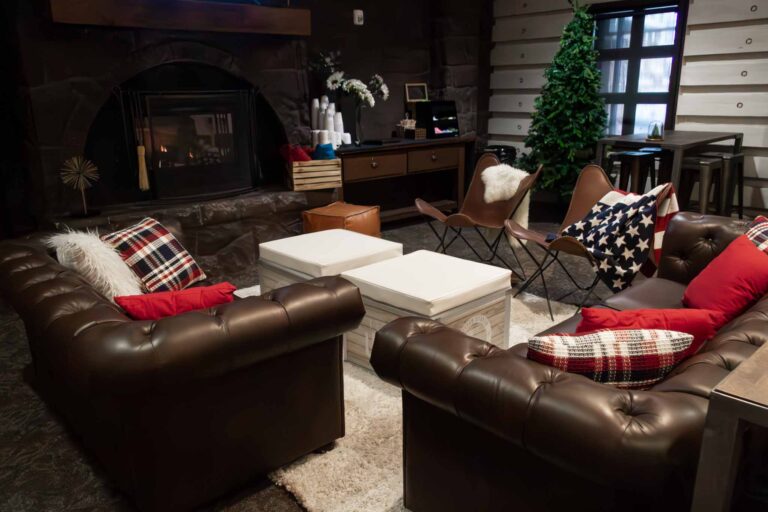 Cozy fireside VIP lounge setup at Park City Mountain featuring brown leather Chesterfield sofas, Team USA branded white ottoman rentals, leather butterfly chairs with faux fur and stars-and-stripes throws, plaid and red accent pillows, a gold starburst decor piece, wooden crate props, and a decorated Christmas tree beside bar-height tables with metal stools.