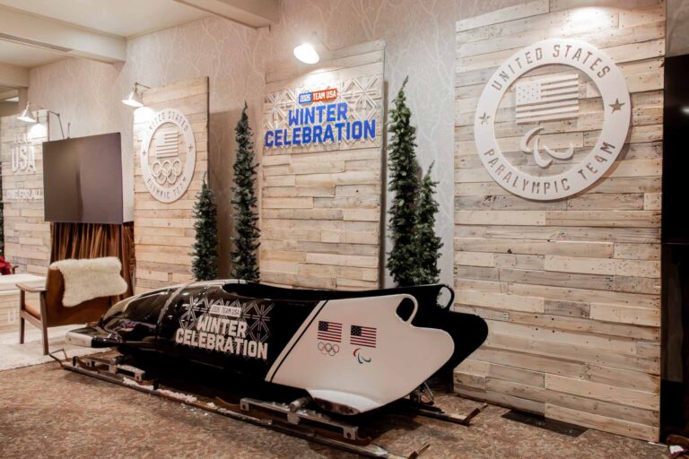 Alt Text: Team USA 2026 Winter Celebration branded bobsled prop displayed in front of a rustic pallet wood accent wall featuring United States Olympic and Paralympic Team medallion signage, flanked by slim decorated Christmas trees at Park City Mountain.