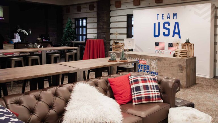 Wide-angle view of the Team USA 2026 Winter Celebration event space at Park City Mountain featuring leather Chesterfield lounge sofas with plaid and faux fur pillows, rustic wood farm tables with metal stools, glass dome centerpieces, a branded reception desk, and a large Team USA backdrop.