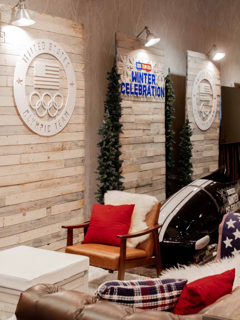Team USA 2026 Winter Celebration lounge area at Park City Mountain featuring United States Olympic Team medallion wall signage on rustic pallet wood panels, slim evergreen trees, a leather armchair with red pillow and faux fur throw, a branded bobsled prop, and layered lounge furniture with plaid and stars-and-stripes accents.