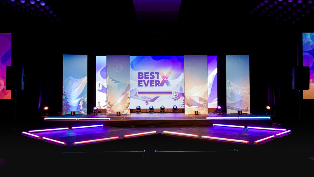 Full front-facing view of the Best Ever X Conference main stage at Hyatt Regency featuring five tall LED screen panels displaying a warm mountain sunrise landscape graphic with the conference logo, neon tube floor lighting in an X formation, moving head stage lighting fixtures, flanking speaker system towers, and purple wash lighting throughout the darkened ballroom.