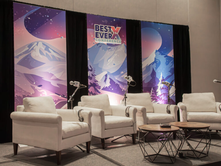 Best Ever X Conference breakout session stage setup at Hyatt Regency featuring four white upholstered armchairs with over-ear headphones, three microphone stands, two round geometric metal coffee tables, and three tall custom printed mountain landscape backdrop panels in pink, purple, and blue with the conference logo, framed by black pipe and drape.