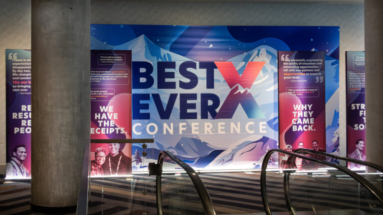 Large-scale Best Ever Conference custom printed backdrop with bold mountain landscape graphics in blue, purple, and red, flanked by tall attendee testimonial banner stands with headshots and quotes, positioned at the top of an escalator entrance at Hyatt Regency.