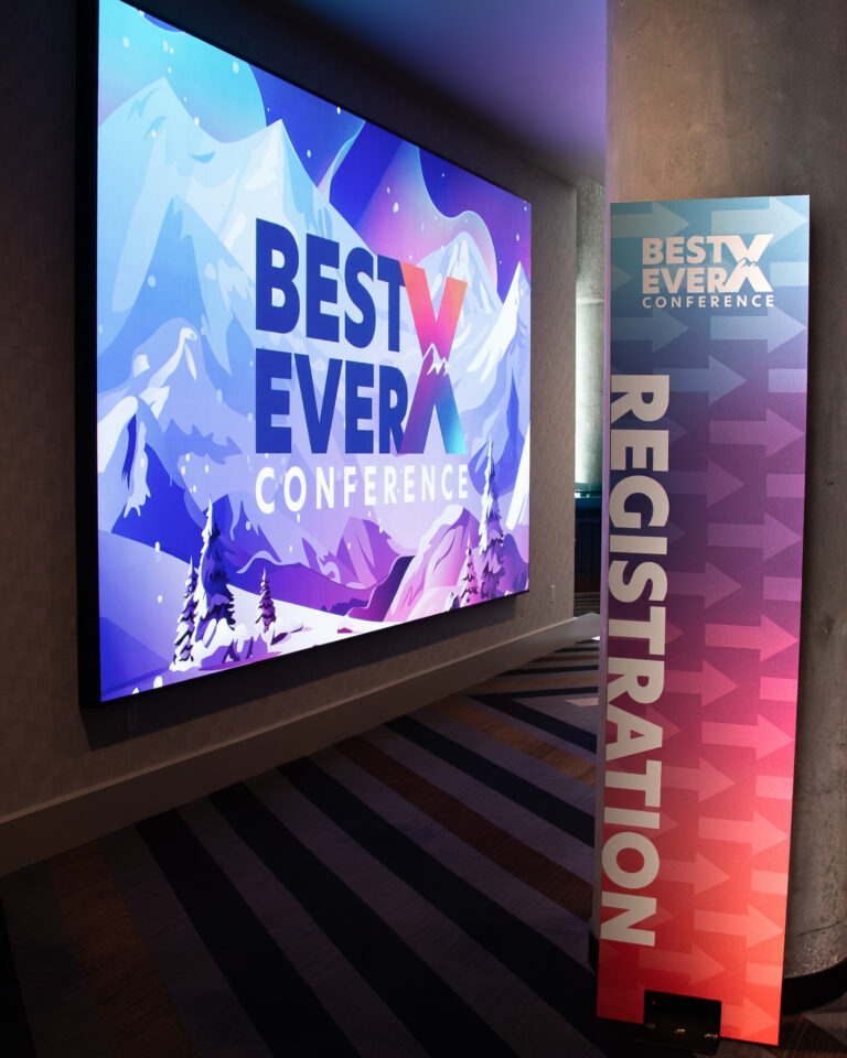 Large flat screen display showing the Best Ever X Conference branded mountain landscape graphic alongside a tall custom printed Registration directional banner stand with gradient purple to pink design and arrow graphics at Hyatt Regency.