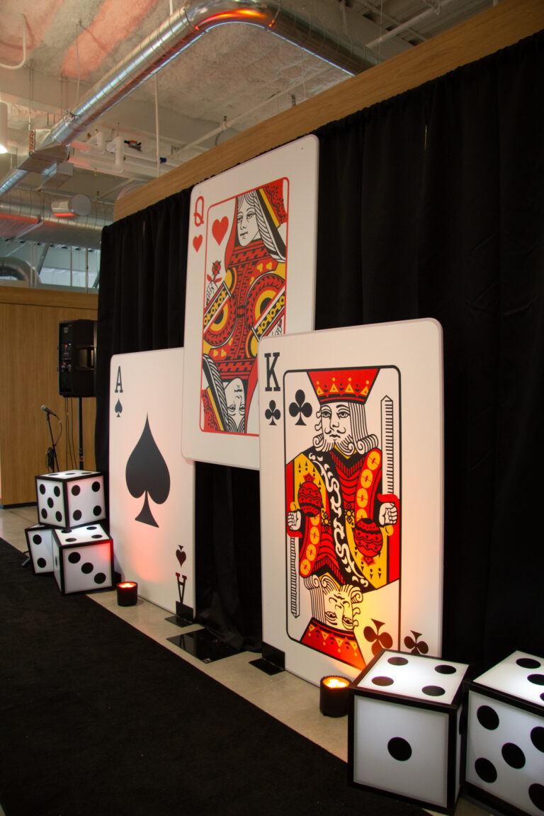 Side-angle view of the Oversized Playing Card Installation showing Ace of Spades, Queen of Hearts, and King of Clubs cards against black pipe and drape with illuminated oversized dice props flanking both sides and red uplighting on ceiling pipes above