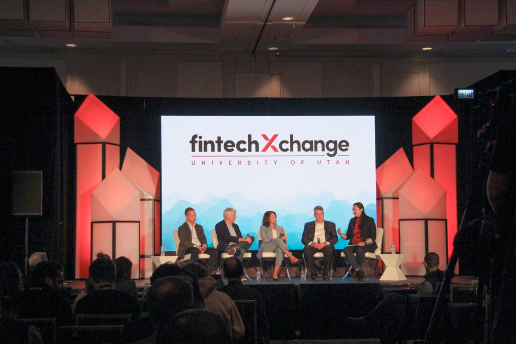 Five panelists seated on a conference stage during a live panel discussion in front of a large LED screen displaying the FintechXchange University of Utah logo, flanked by red-lit Shard Tower scenic structures and a seated audience in the foreground