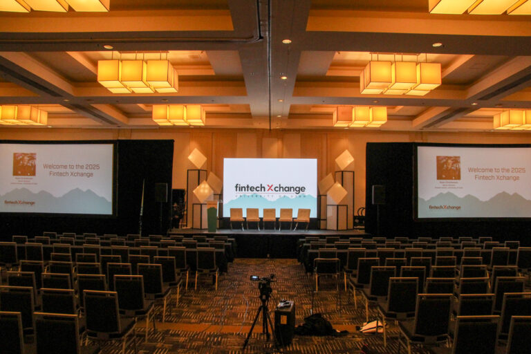 Wide view of a fully dressed conference ballroom with a center LED screen displaying the FintechXchange logo, Shard Tower scenic structures flanking the stage, dual projection screens, panel seating, rows of audience chairs, and a camera on a tripod in the center aisle