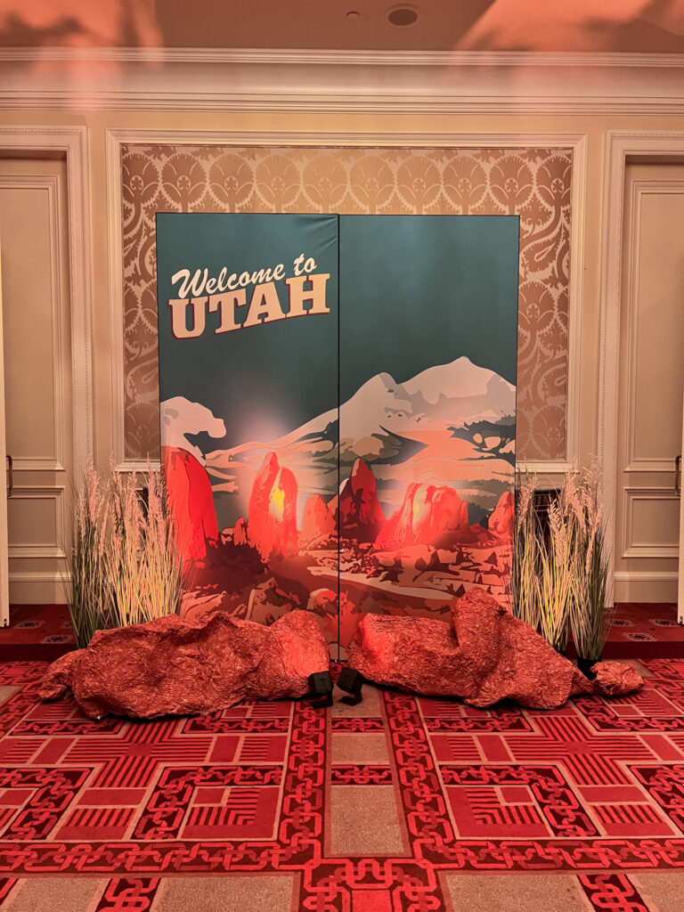 Close-up of Welcome to Utah illustrated step and repeat backdrop with vintage travel poster-style red rock graphic, faux sandstone boulder props, desert grass arrangements, and saturated red uplighting at The Grand America Hotel Salt Lake City for 360 Destination Group corporate event