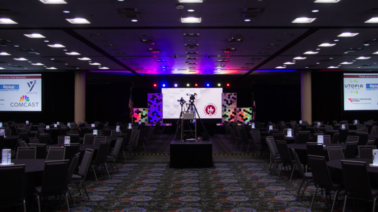 A large ballroom at the Salt Palace set with round banquet tables, a main stage featuring hexagonal-patterned backdrops, and large projection screens displaying gold sponsor logos like Comcast and Utopia Fiber.