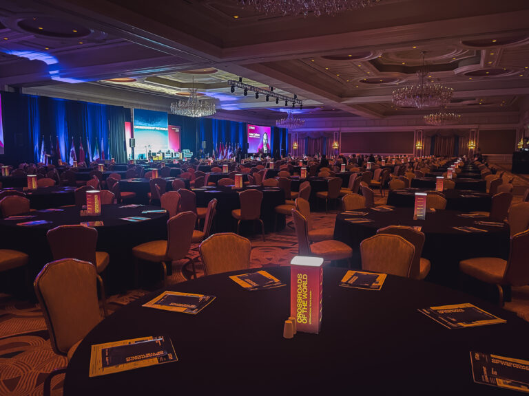 Conference ballroom setup with a custom stage, large projection screens, international flags, and branded illuminated centerpieces at the Crossroads of the World International Trade Summit.