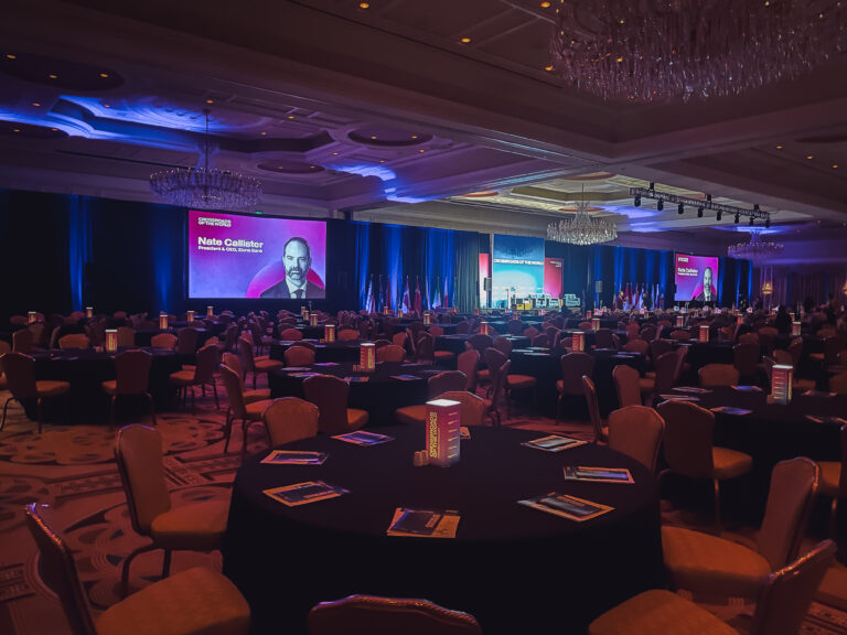 Large conference ballroom with custom stage production, dual presentation screens, international flags, and branded illuminated centerpieces at the Crossroads of the World International Trade Summit.