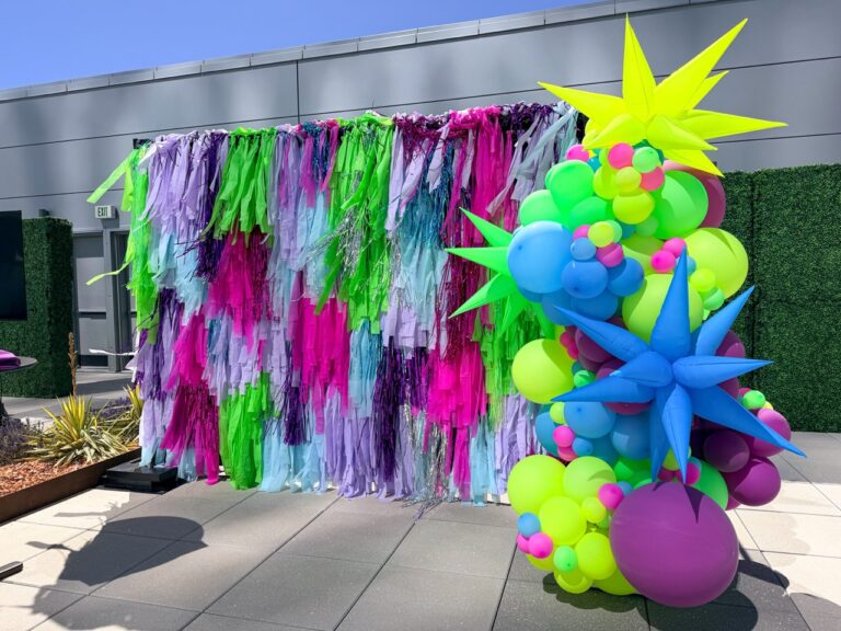Vibrant outdoor event photo backdrop featuring neon fringe draping and a colorful balloon installation for a corporate event activation.