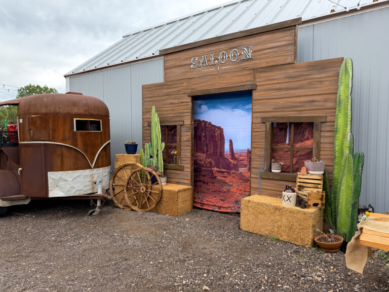 Western saloon facade with a vintage horse trailer, wagon wheels, hay bales, cacti, and rustic decor at The Lodge at Blue Sky.