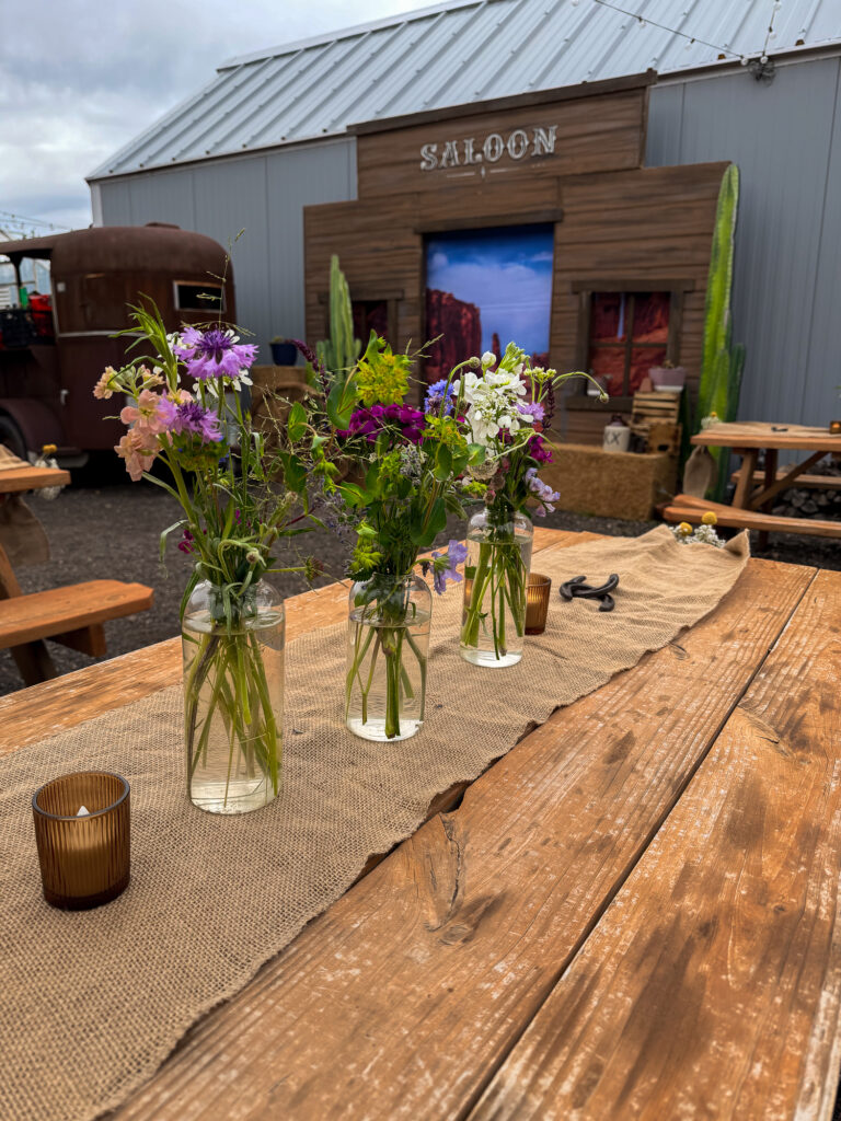 Rustic picnic tables styled with wildflower centerpieces, burlap runners, candles, and a Western saloon backdrop at The Lodge at Blue Sky.