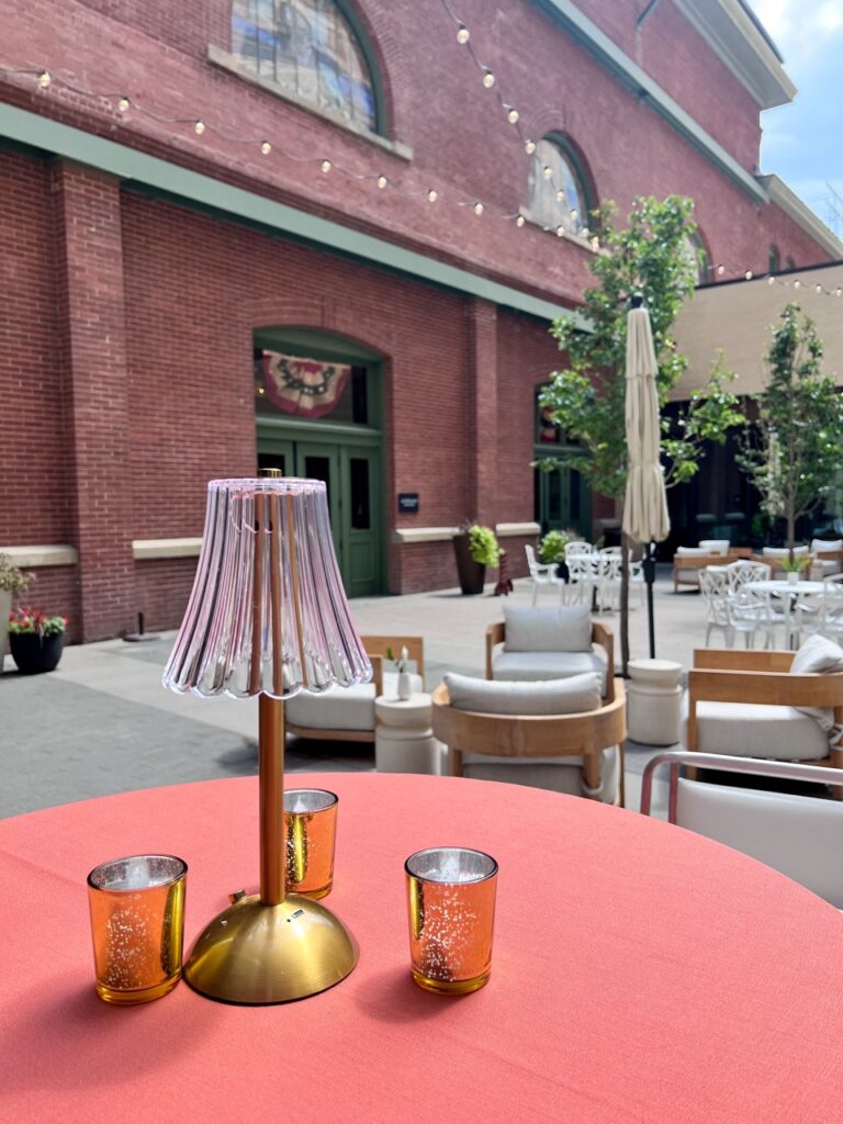 Outdoor cocktail table featuring a coral linen, wireless LED table lamp, and mercury glass votive candles in the courtyard of Asher Adams Salt Lake City.